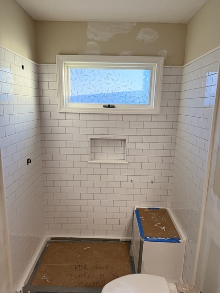 Bathroom remodel in progress: white subway tile walls, a niche shelf, window with patterned privacy film, and a toilet covered with cardboard. The shower floor remains unfinished—an interesting case study from Columbia SC.