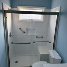 A modern bathroom remodel in Columbia SC features a glass-enclosed shower with white tile, safety grab bars, a built-in bench, recessed shelf, frosted window, and a toilet next to a sleek white countertop.