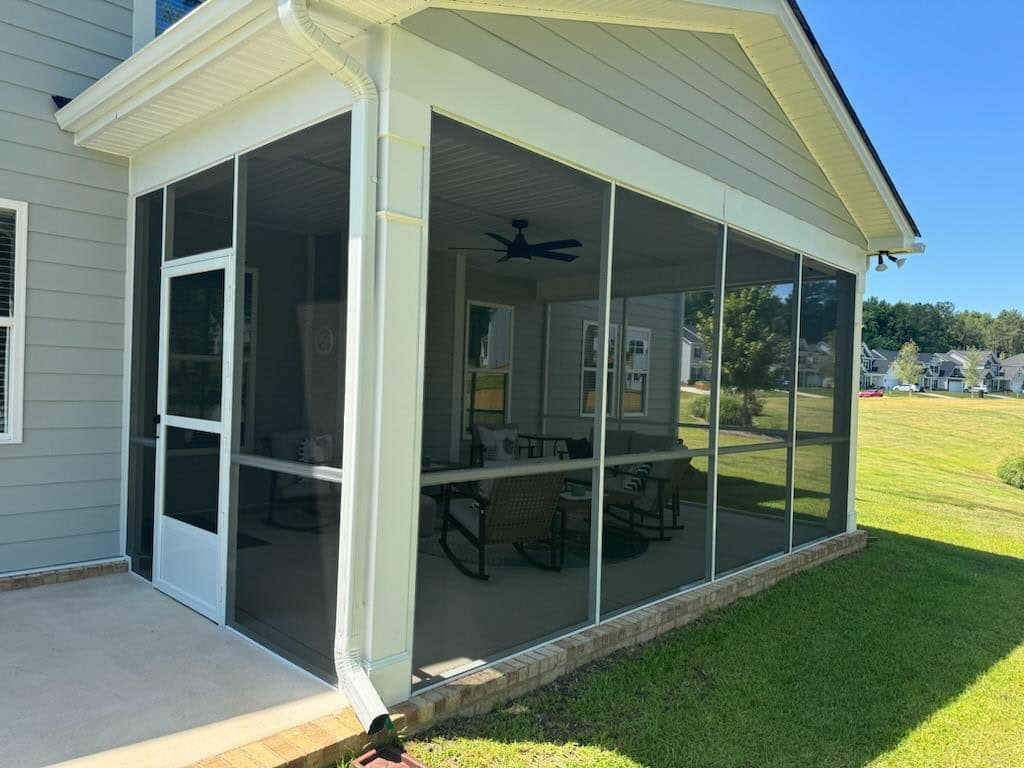 A custom screen porch attached to a house with light gray siding, featuring several chairs and a ceiling fan inside. The porch overlooks a green lawn with neighboring houses in the background on a sunny Columbia, SC day.